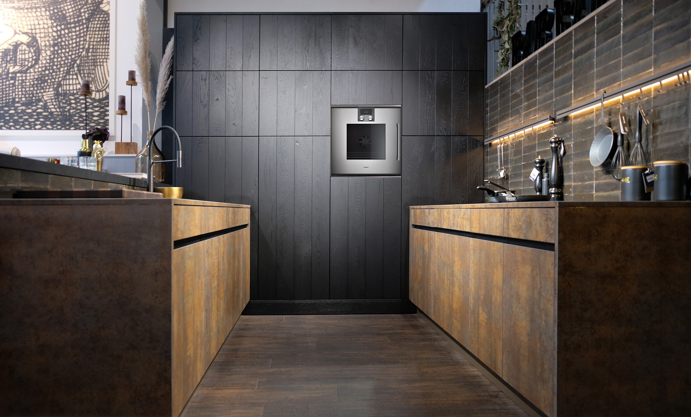 Zwei gegenüberliegende Küchenzeilen ohne Oberschränke. Am Ende mit eingebauten Schränken in einer dunklen Holzstruktur. Dazu ein hochgebauter Backofen in schlichtem Design. Die Unterschränke haben eine metallische Oberfläche in Kupferfarben. Die Küche ist mit Griffleisten ausgestattet, grifflos. Die Seite links ist mit einer angebauten Tresenplatte in dunklem Massivholz versehen. Die rechte Seite steht vor einer Wand mit schwarzer Leiste, die über Haken und Beleuchtung verfügt. Die Küche ist im Hummel Küchenwerk City Store in Hamburg zu besichtigen.