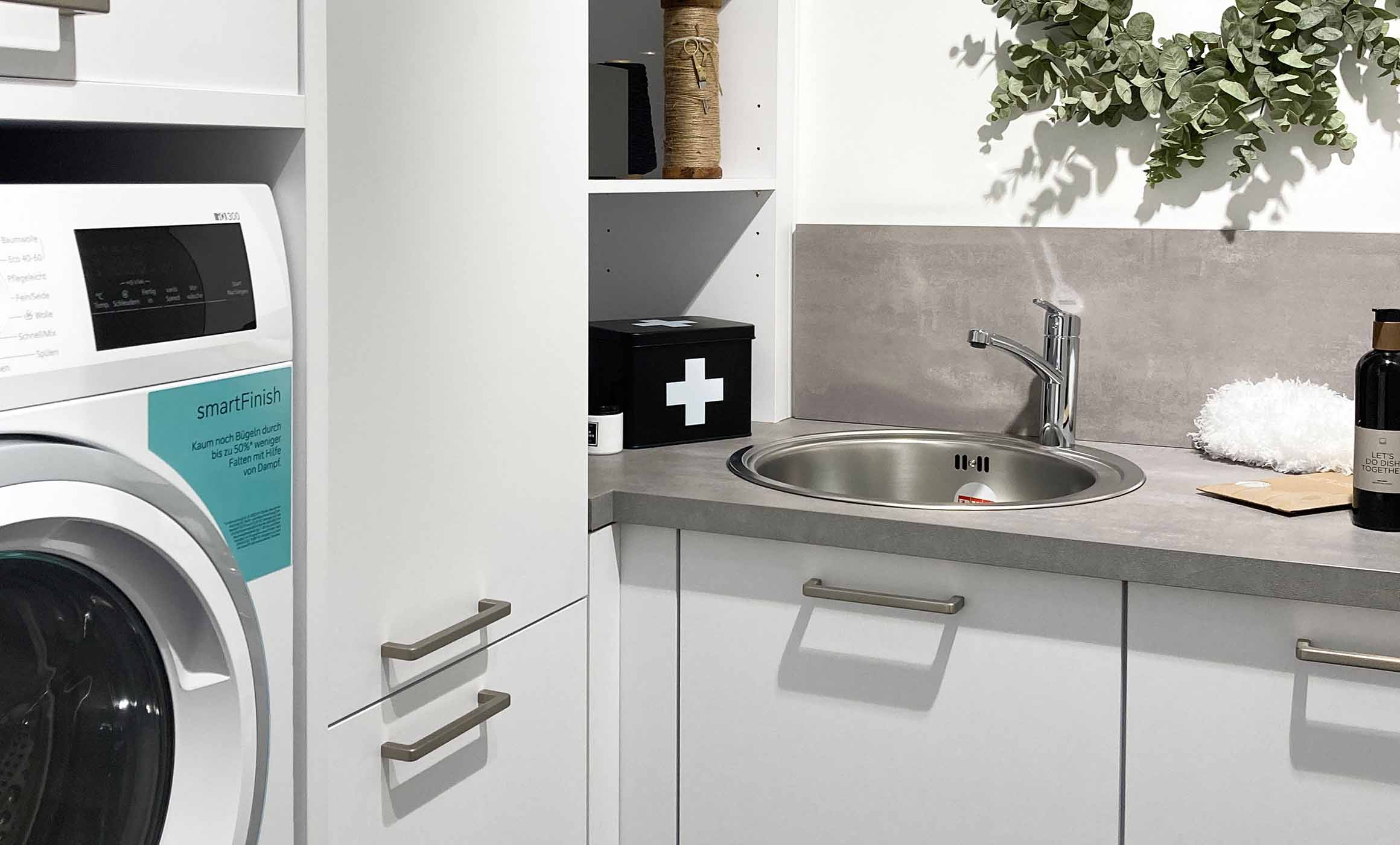 Hauswirtschaftsraum in weiß mit Unterschränken und Hochschränken mit Küchenmöbeln geplant. In L-Form mit einer Arbeitsplatte in Betonoptik. Der Hochschrank auf der linken Seite beherbergt die Waschmaschine und Körber für die Schmutzwäsche. Die Waschküche bietet viel Stauraum und verbirgt sich hinter einer Küchenfront, die in der Küche als Tür fungiert. Zu sehen im Hummel Küchenwerk Norderstedt.
