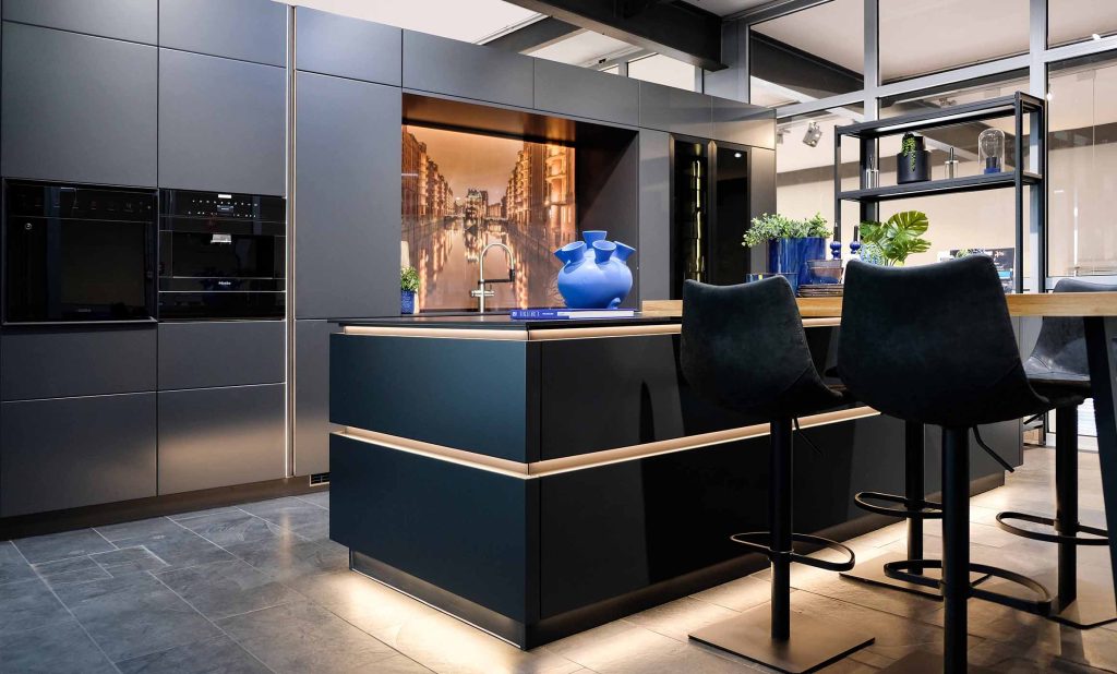 Diese Küche wirkt modern und elegant mit dunklen, glatten Fronten. Die Arbeitsfläche der Kücheninsel ist mittig angeordnet, mit Barhockern davor, beleuchtet durch indirekte LED-Beleuchtung im Sockel. Hochwertige Einbaugeräte sind sauber in die Schrankwand integriert. Dekorationen wie blaue Vasen und grüne Pflanzen setzen farbliche Akzente. Große Fenster sorgen für viel Tageslicht und eine offene, freundliche Atmosphäre.