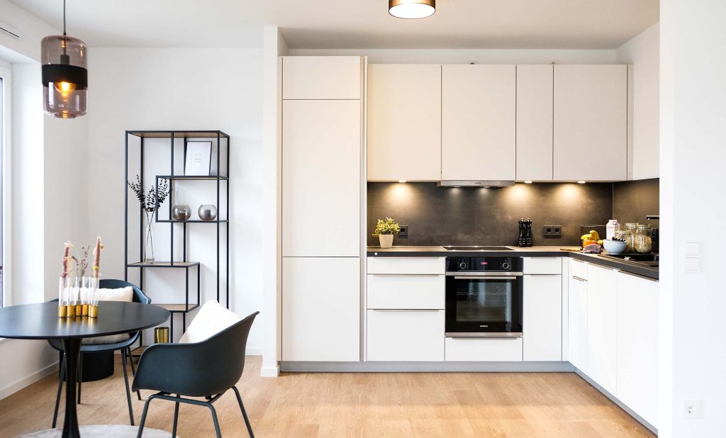 Man blickt in eine offene Wohnküche. Links befindet sich der runde, schwarze Essplatz mit schwarzen Stühlen dazu. An der Wand steht ein Metallregal mit Dekoelementen. Recht ist eine weiße Küche im L-Format mit dunkler Arbeitsplatte und Nischenverkleidung eingebaut. Jeweils an den Außenkanten befinden sich Hochschränke. Dazwischen sieht man Unterschränke mit Auszügen und Oberschränke, die mit Höhe der Hochschränke abschließen. Die Küche ist beleuchtet und mit Küchenutensilien dekoriert.
