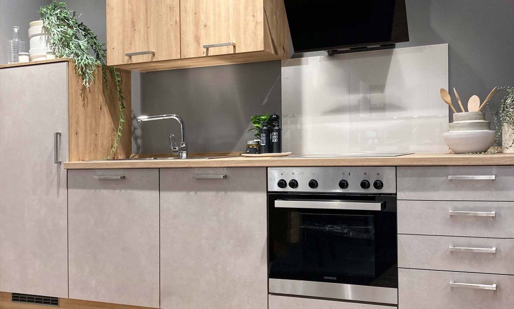 Zu sehen ist eine kompakte Küchenzeile mit einem 3/4 hohen Schrank links. Daneben sind Ober- und Unterschränke angeordnet. Ein Backofen und darüber eine schwarze Dunstabzugshaube. Der Spritzschutz hinter dem Herd ist in einer hellen Farbe. Die Front der Oberschränke ist aus hellem Holz. Die Unterschränke sind grau-meliert. Die Farbkombination wirkt warm.