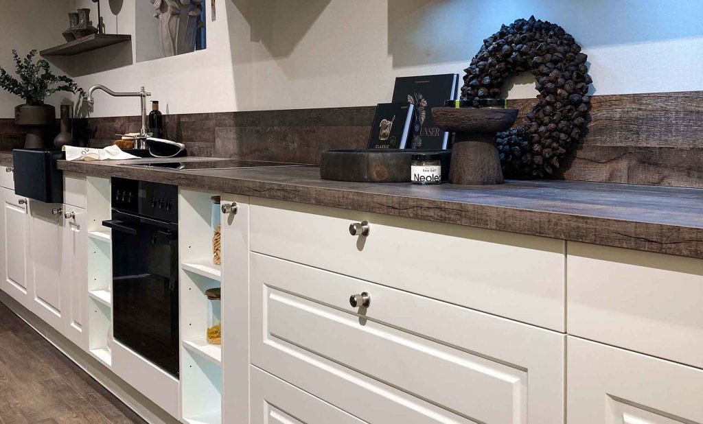 Das Bild zeigt eine moderne, helle Küche im Landhausstil mit cremefarbenen Unterschränken und Auszügen. Die Arbeitsplatte und der Spritzschutz sind aus dunkelbraunem Holz. Es gibt einen schwarzen Backofen, offene Regale mit Vorratsgläsern und eine Spüle mit Armatur im Retrolook. Dekorationen wie ein Kranz, Bücher und Vasen stehen auf der Arbeitsfläche und im Regal. Die Küche wirkt gemütich.