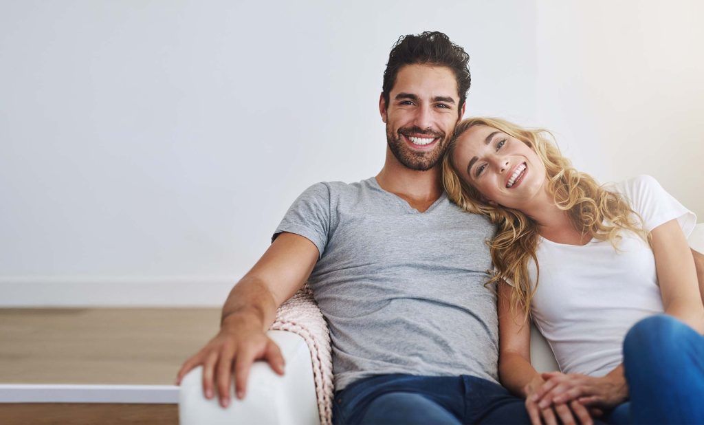 Junges Paar auf dem Sofa. Lächeln freundlich in die Kamera. Der Raum drumherum ist leer.
