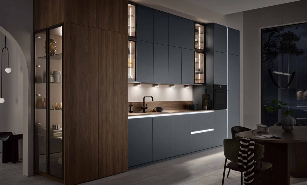 Die gezeigte Küche ist als Zeile mit seitlichem Abschluss in Form einer beleuchteten Vitrine auf der linken Seite geplant. Die Hochschränke auf der linken Seite bestehen aus dunklem Holz, wie auch die Arbeitsfläche. Unterschränke, Oberschränke und seitliche Hochschränke (links) in einem mittleren Blaugrau-Farbton. Die Unterschränke sowie die Hochschränke haben beleuchtete Griffleisten. Zur Auflockerung sind im Oberschrankbereich Vitrinen mit Klarglas verbaut. Die offene Küche wird in einer abendlichen Stimmung dargestellt. Vor der Küchenzeile ist ein Esstisch in dunklem Holz platziert.
