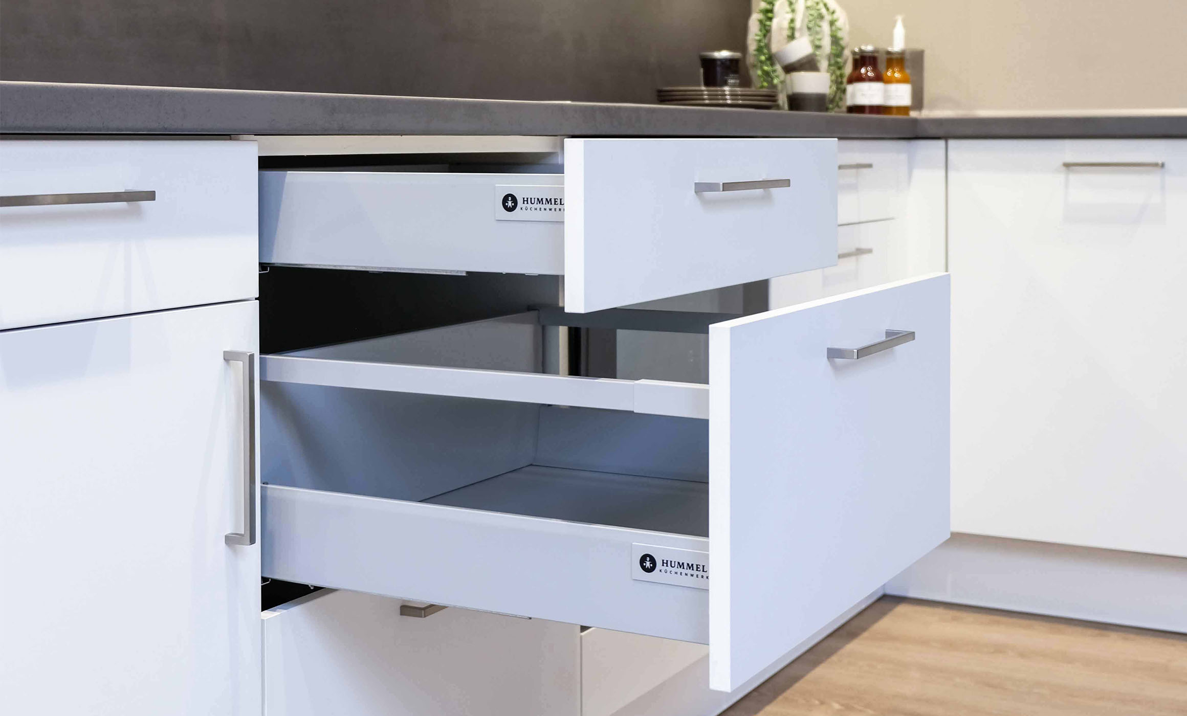Ein aufgezogener Auszugschrank einer Küche, mit leeren Auszügen und dem Logo von Hummel ist zu sehen. Farbe der Front weiß, Arbeitsplatte in grau.