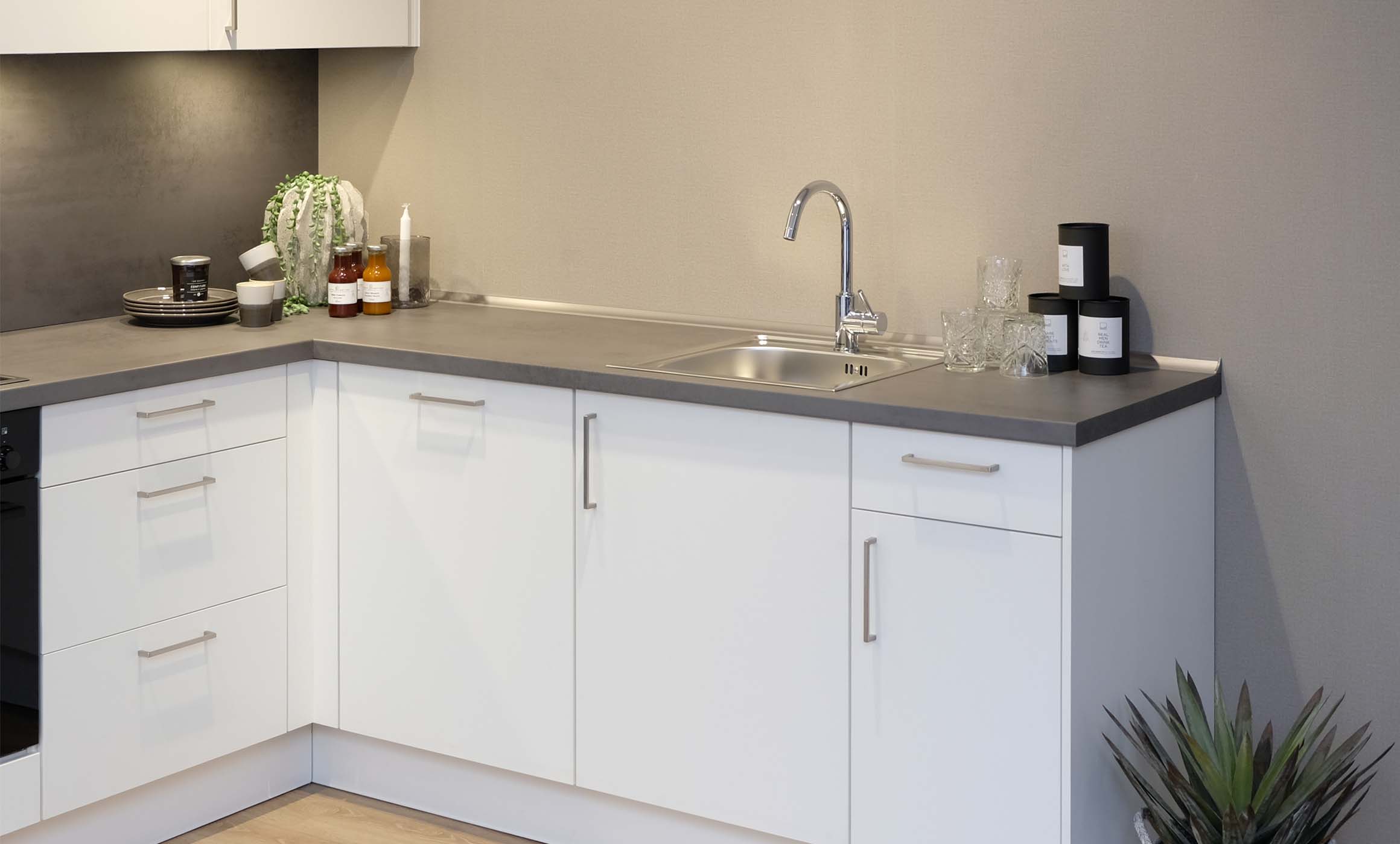 Unterschränke einer Küche in Weiß mit einer grauen Arbeitsplatte. Eine Spüle in Edelstahl mit chrom Wasserhahn sowie ein paar Gläser und Dosen in Schwarz, sind zu sehen.