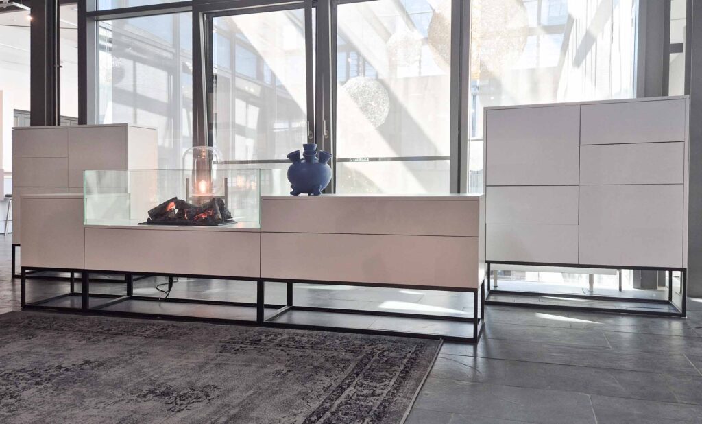 Sideboard in weiß mit Push-to-open Front. Gestell schwarz als Kubus mit Vierkantstreben. Wirkt leicht. In der Mitte befindet sich ein gläserner Kasten, in dem ein künstliches Feuer leuchtet.