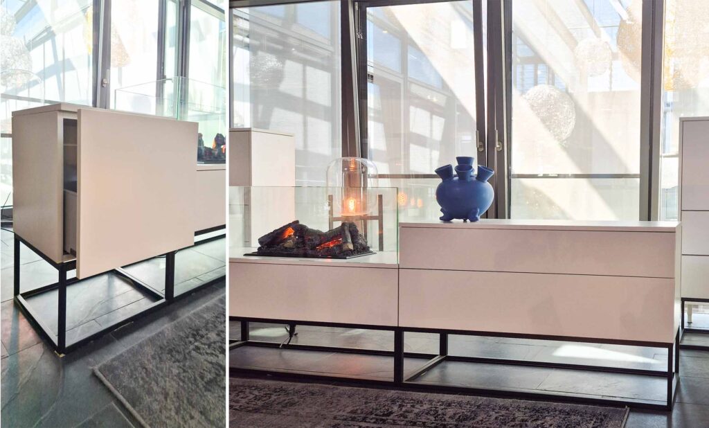 Sideboard in weiß mit Push-to-open Front. Gestell schwarz als Kubus mit Vierkantstreben. Wirkt leicht. In der Mitte befindet sich ein gläserner Kasten, in dem ein künstliches Feuer leuchtet.