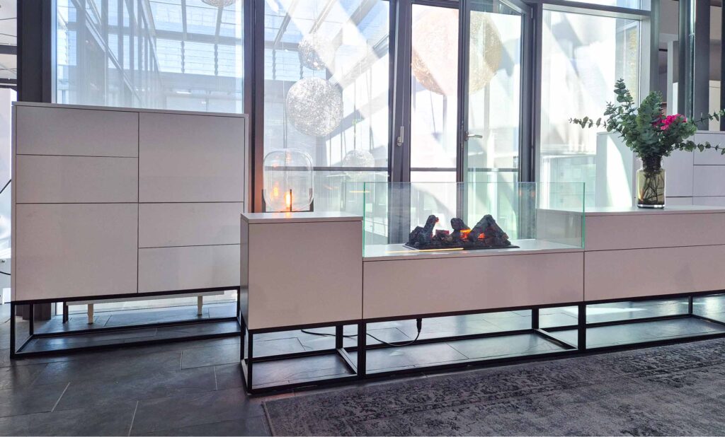 Sideboard in weiß mit Push-to-open Front. Gestell schwarz als Kubus mit Vierkantstreben. Wirkt leicht. In der Mitte befindet sich ein gläserner Kasten, in dem ein künstliches Feuer leuchtet.
